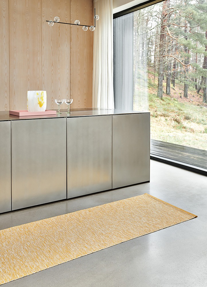 A contemporary kitchen interior featuring the Pappelina Sam mustard rug, placed on a polished concrete floor. The room includes a metal kitchen island with decorative glassware and a light wood paneling, and large windows with a view of the forest.